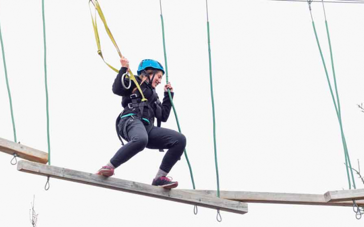 A person wearing safety gear and secured by ropes balances on an obstacle on a high ropes course. 